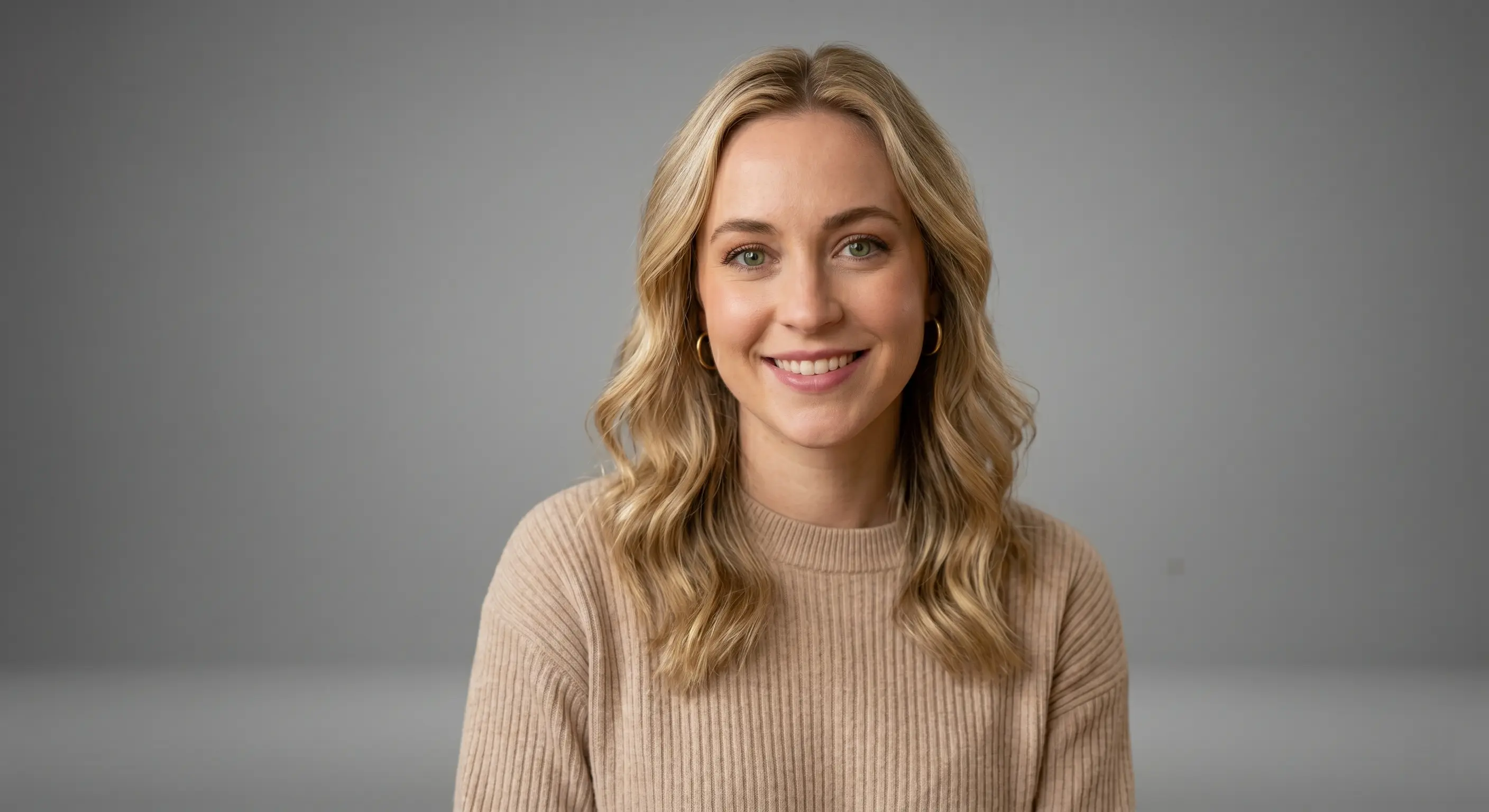 Blonde woman with long wavy hair wearing a beige ribbed knit sweater, smiling in a headshot against a plain grey studio backdrop