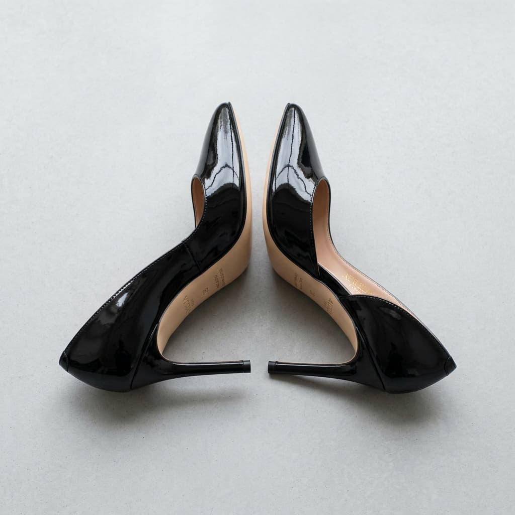 Pair of black patent stiletto heels with nude soles, shot from behind on a pale grey surface